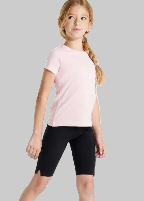 A young girl with a blonde braid wearing a light pink short-sleeve t-shirt and black bike shorts, standing against a plain light gray studio background.