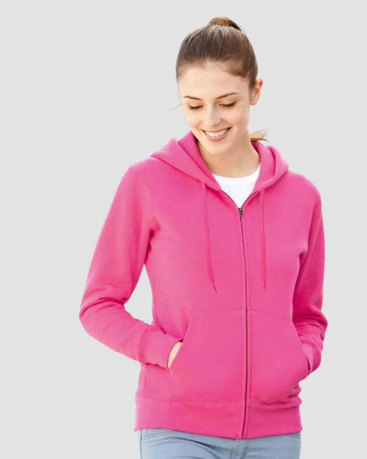 A woman wearing a bright pink zip-up hoodie over a white t-shirt and light blue jeans, standing outdoors against a white wall with a blue base.