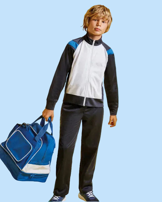 A child model wearing a colorblock athletic tracksuit with a white, navy, and black jacket and matching pants, holding a blue and white duffel bag against a solid bright cyan background.