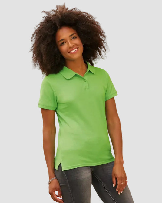 A woman with dark curly hair wearing a bright lime green short-sleeve polo shirt and dark gray jeans, standing outdoors in front of a beige stone building.