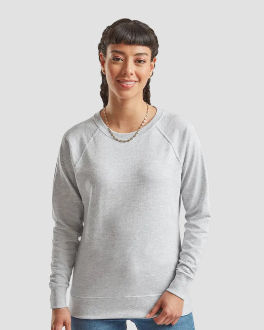A woman wearing a light gray heather crewneck sweatshirt with raglan sleeves, paired with blue jeans and a gold chain necklace, against a solid light gray background.