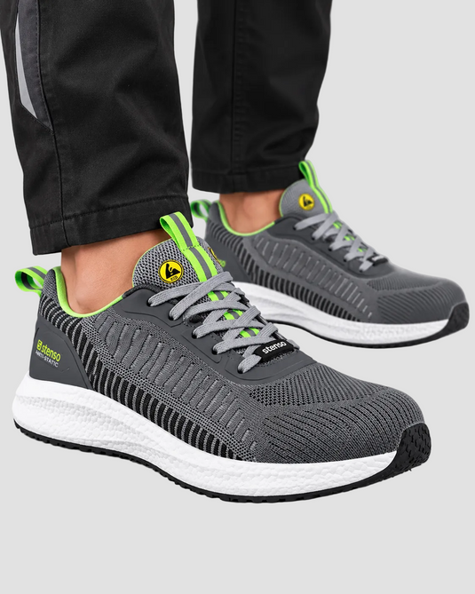 A pair of gray running shoes with bright neon green accents and white midsoles, shown on a model wearing black pants against a light gray studio background.