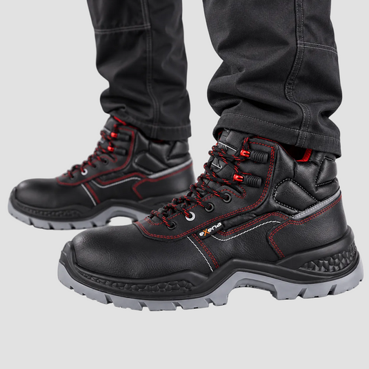 Black leather safety work boots with red contrast stitching and red accents on the laces, shown on a person wearing dark gray pants against a light gray studio background.