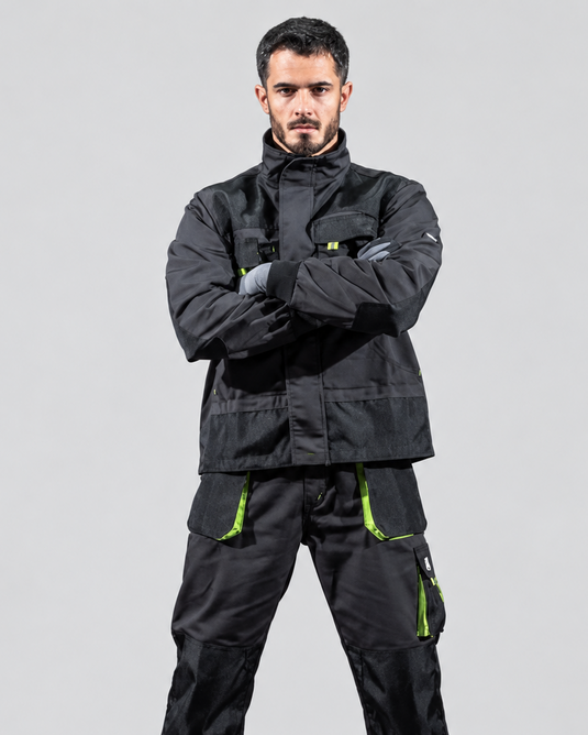 A man modeling a black technical workwear set consisting of a jacket and pants with bright green zipper accents, photographed against a neutral gray studio background.