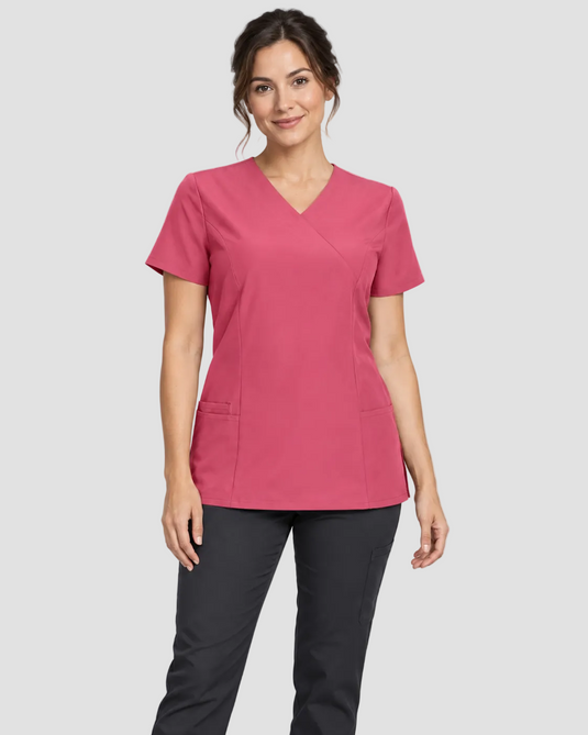 A woman modeling a short-sleeve V-neck scrub top in dusty rose pink, paired with dark gray scrub pants, against a light gray studio background.